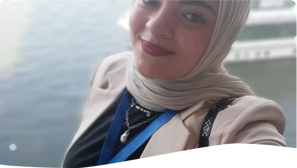 A smiling woman wearing a beige blazer and a light-colored hijab, with sunglasses resting on her head. She has a lanyard around her neck and is taking a selfie in front of a large body of water, with blurred boats in the background